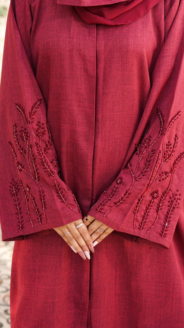 A maroon abaya made from soft, textured linen fabric, featuring elegant embroidery on the sleeves and a modest, flowing silhouette.